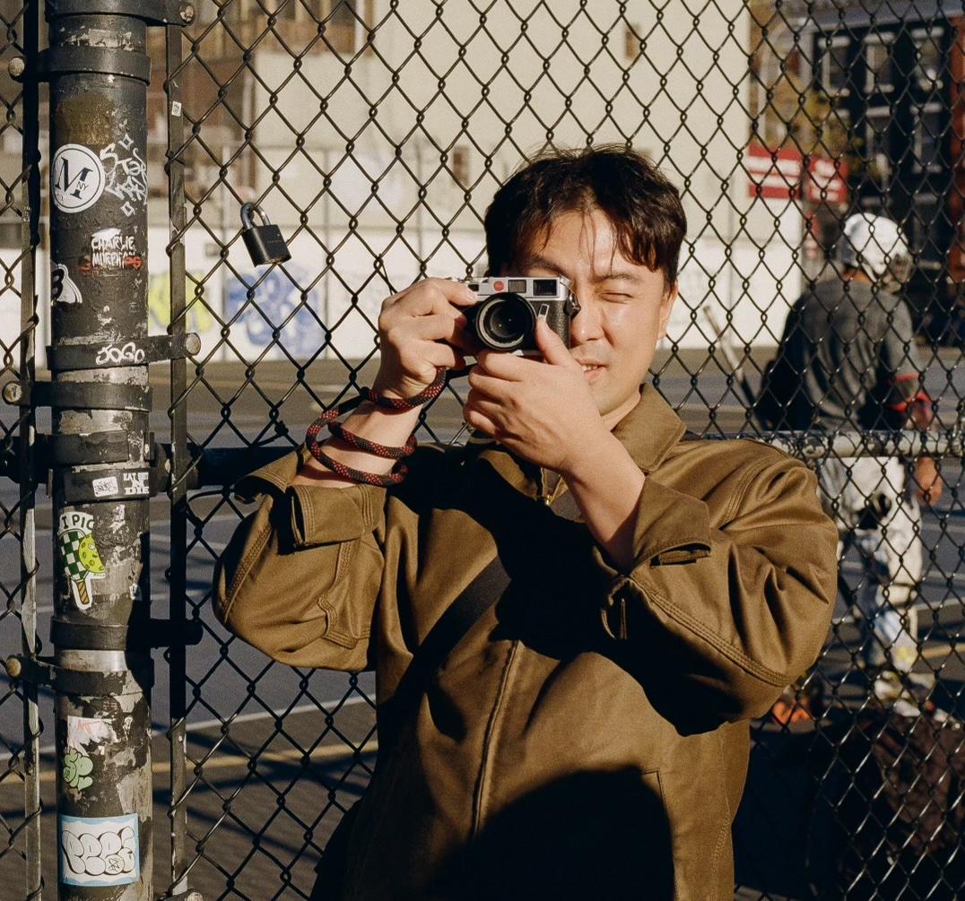 David Lee raising a camera to his eye in front of a chain-link fence